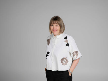 Person wearing a white shirt with abstract patterns and black trousers, standing against a grey background.