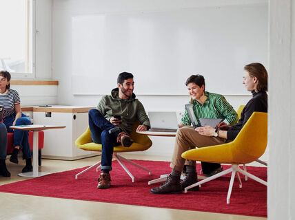 Viisi ihmistä istuu modernissa toimistotilassa kannettavien tietokoneiden ja muistikirjojen kanssa keskustelemassa. Punainen matto ja värikkäitä huonekaluja.