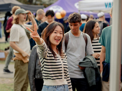 A person wearing a striped shirt makes a peace sign at an outdoor event with people in the background.