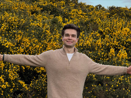 Person in beige jumper stands arms outstretched in front of dense yellow-flowered bushes outdoors