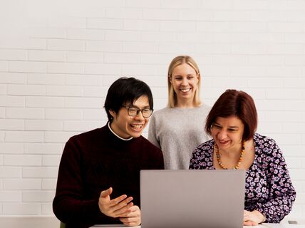 Three people working together in front of a laptop, against a white brick wall background.