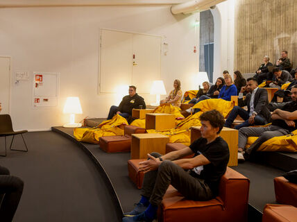 En person med mikrofon talar till en publik som sitter på saccosäckar och stolar. En presentationsskärm syns.