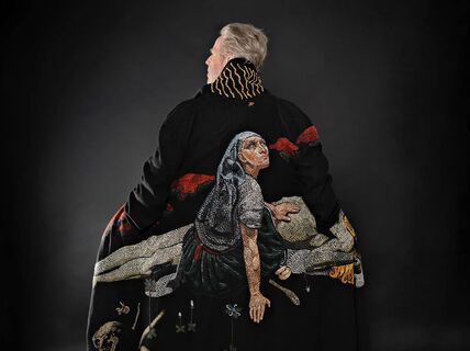 Person from behind in dark coat with large embroidered scene of kneeling figure on dramatic black background