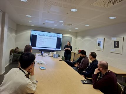 A meeting room with a presentation on a screen. Six people are seated at a table facing a presenter.