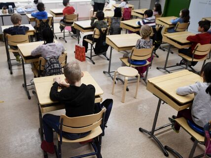 Primary pupils sit spaced at wooden desks in a bright classroom, facing the teacher at the front.