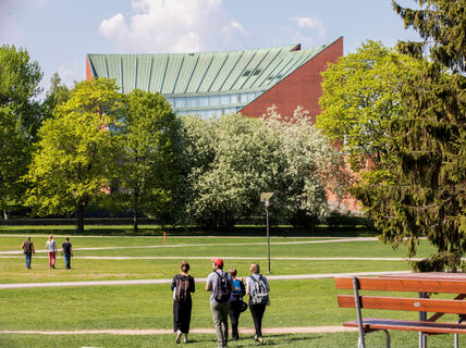 Opiskelijat kävelevät aurinkoisella kampuksen nurmella, taustalla punatiilinen rakennus ja puita