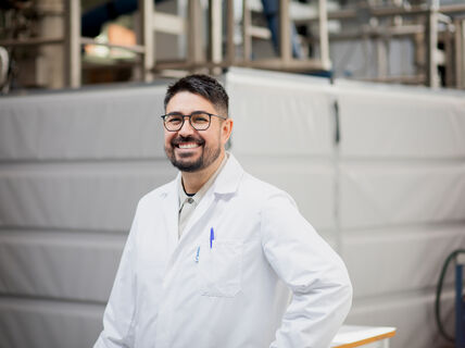 Person in a white lab coat stands in an industrial facility, smiling, blue pen in pocket.