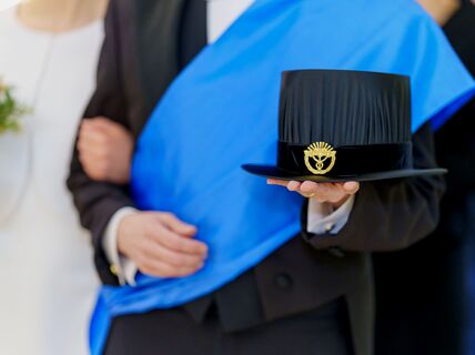 Person in formal suit with blue sash holding a black doctoral hat with a gold emblem