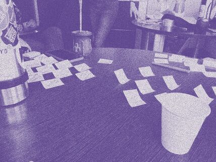 Workshop photo digitally modified with a purple duotone effect. The picture shows a table with sticky notes, a pen, a phone, a cup, thermos and mate, with people and other table in the background.