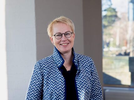 Smiling person with short blond hair and glasses in a zigzag blue-white jacket, standing indoors by a window