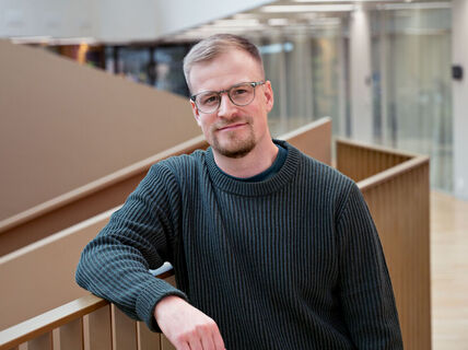 Man with glasses and short beard in dark jumper, leaning on railing in bright modern office