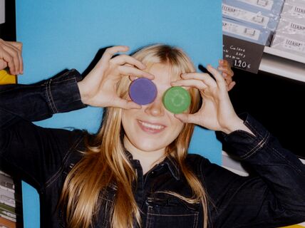 Smiling blonde person holds purple and green discs over her eyes, posing playfully in a colourful shop.