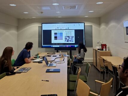Four people in a meeting room watch a large screen showing charts and photos, with laptops on a long table.