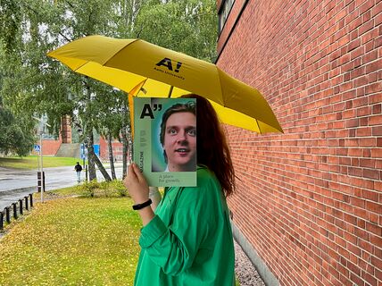 Person in green holds magazine over face under yellow Aalto umbrella by wet brick wall and trees
