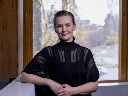 Woman in a black top leaning on a white railing indoors, large windows showing a blurry autumn cityscape