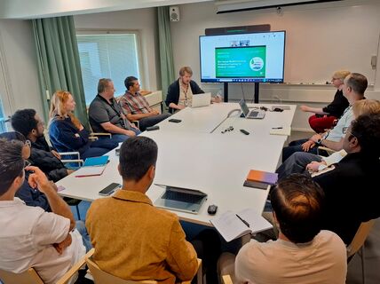 People seated around a white table in a meeting, watching a laptop presentation on a large screen.
