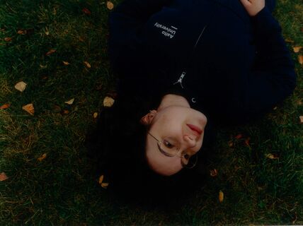 Person in glasses lying on green grass with autumn leaves, wearing a dark Aalto University hoodie