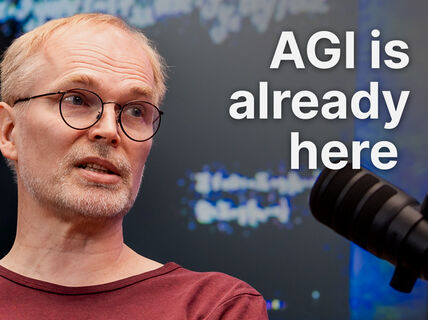 Middle‑aged man in glasses speaks into a black studio microphone, text reads “AGI is already here”.