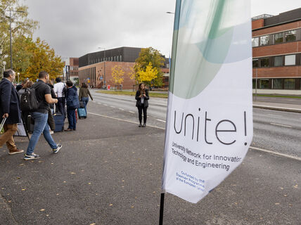 Ihmiset matkatavaroiden kanssa odottavat kampustien varrella Unite!-lipun ja syyspuiden lähellä.