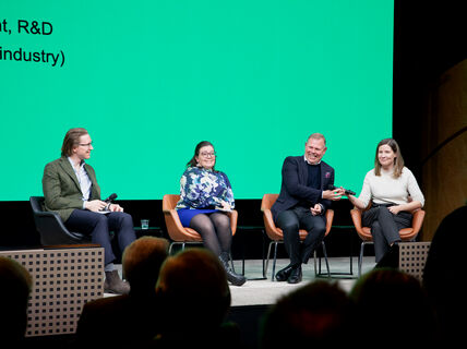 Neljä paneelikeskustelijaa istuu lavalla mikrofonien kanssa, vihreä tausta