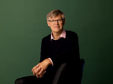 Middle‑aged man with glasses seated, wearing dark jumper and pink shirt, against plain green background.