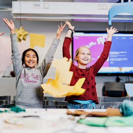 Three children with arms raised in a classroom, surrounded by colourful paper crafts and a monitor in the background.