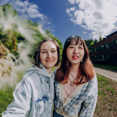 Kaksi ihmistä poseeraa ulkona, taustalla vihreitä puita ja rakennus, etualalla voikukka.