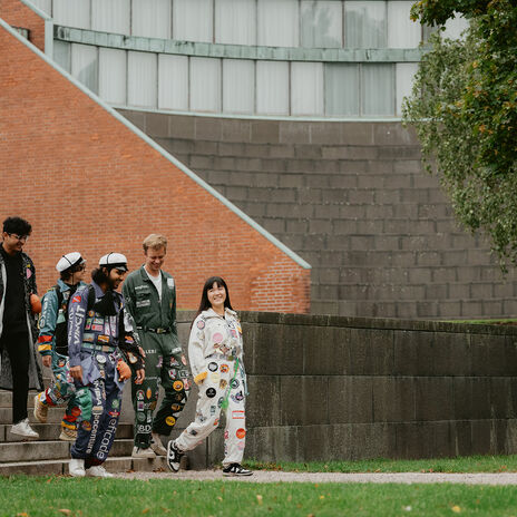A group of people wearing colourful student overalls walk down steps beside a brick building and a large tree.