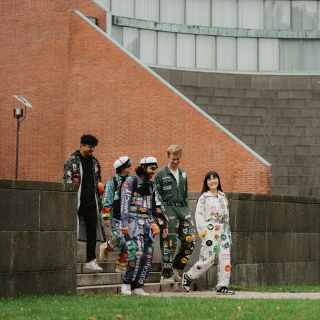 A group of people wearing colourful student overalls walk down steps beside a brick building and a large tree.