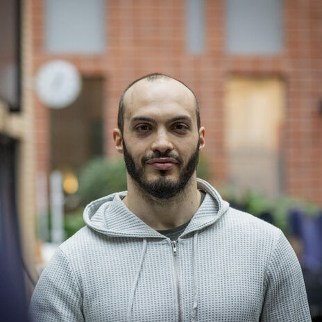 A person wearing a light grey hoodie stands indoors with a brick wall and green plants in the background.