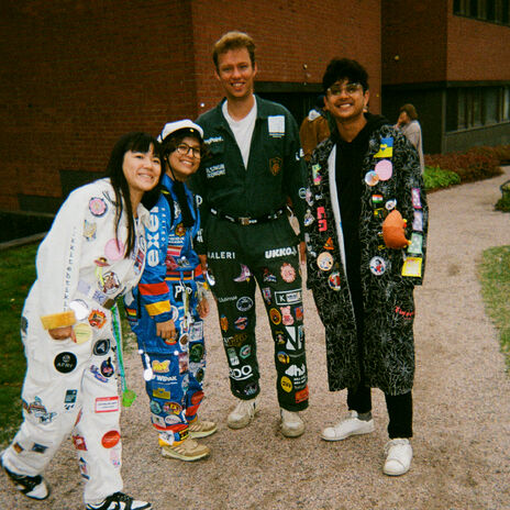 Neljä ihmistä värikkäissä opiskelijahaalareissa, joissa on paljon merkkejä, seisoo tiilirakennuksen ulkopuolella.