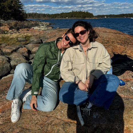 Two people sitting on rocky shore with sea and trees in background. One wears green jacket, other beige jacket.