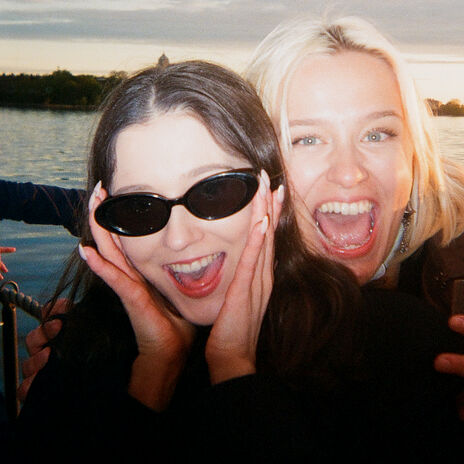 Kaksi ihmistä veneessä, taustalla vettä ja puita. Toisella on tumma tukka, toisella vaalea tukka.