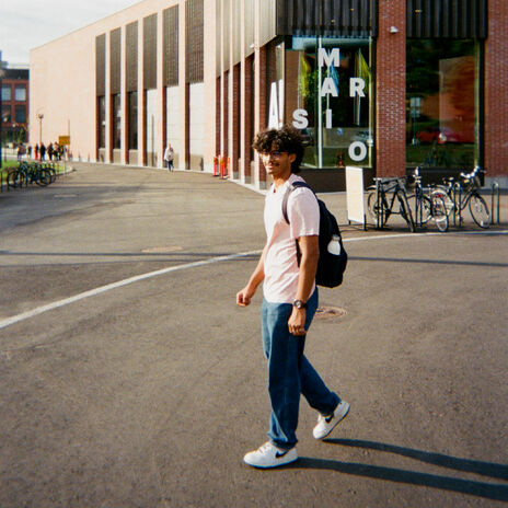 A person in a pink shirt and jeans walks near a modern building with large windows and bicycles parked outside.
