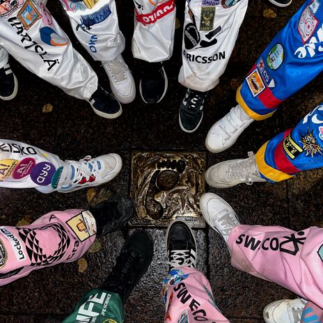 A group of people in colourful overalls with patches stand in a circle around a bronze plaque with a footprint.