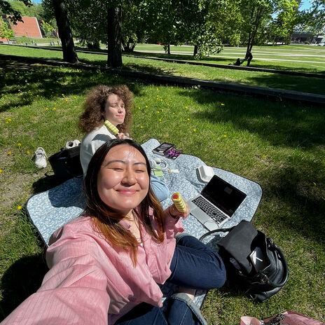 Two people having a picnic on a blanket with a laptop, bags, and food in a park with trees and grass.