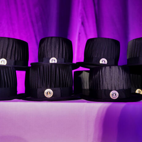 En rad svarta akademiska hattar med silveremblem på ett bord, mot en lila bakgrund.