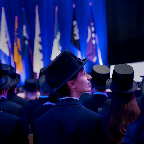 Graduates in dark suits and top hats at a ceremony, facing a blue-lit stage with many flags.