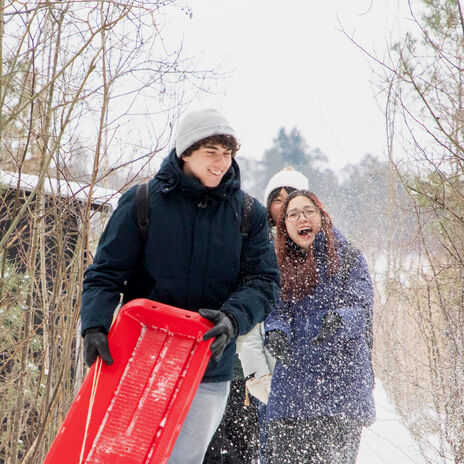 People walking in snowy woods, one holding a red sled. One person is throwing snow in the air.