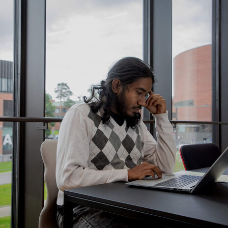 A person wearing a white and grey argyle sweater, sitting at a table with a laptop in front of them, near large windows.