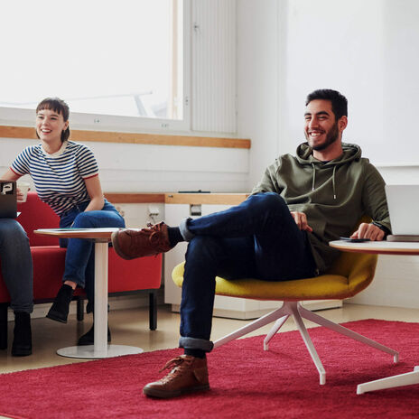 Kaksi ihmistä istuu sohvilla ikkunan edessä keskustellen. Kolmas henkilö istuu etualalla keltaisella nojatuolilla, hän on ristinyt jalkansa ja näyttää nauravan.