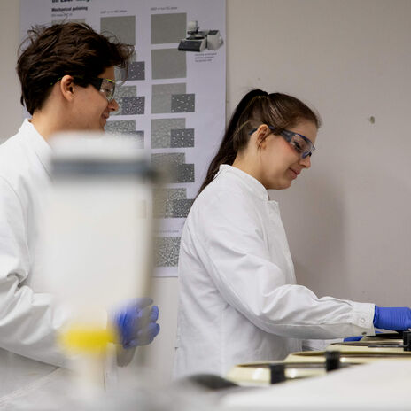 Två laboratoriearbetare i vita rockar och blå handskar arbetar vid bänkutrustning