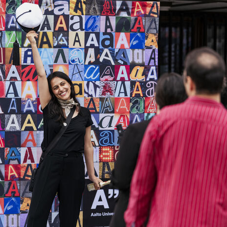 Person in black raising white cap before crowd, standing by colourful Aalto University letter A collage wall