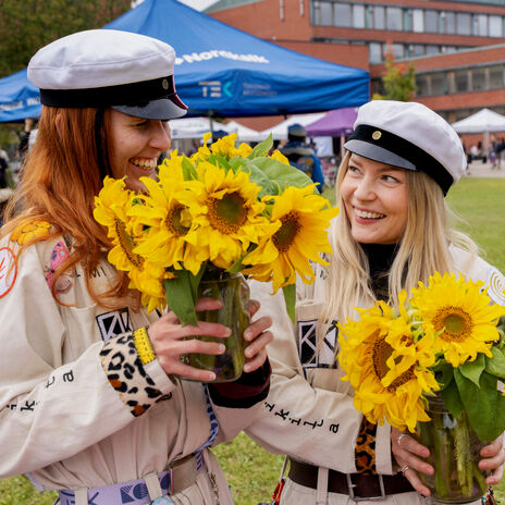 Two smiling students in white caps on a lawn, holding bright yellow sunflowers in glass jars at an outdoor event