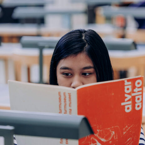 Student with long dark hair reading an orange and white Alvar Aalto book in an empty classroom