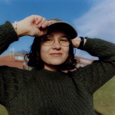 Person in green jumper and cap smiling outdoors, holding cap brim with both hands on a sunny day