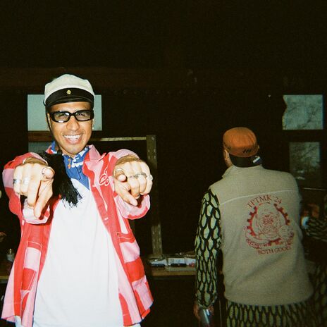 Smiling person in sailor cap and red jacket pointing at camera, with others talking in dim indoor space