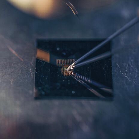 Close-up of thin probes testing a small gold microchip on a dark metallic surface