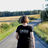 A woman is walking on a gravel road in a summer field landscape, away from the camera. Her black t-shirt reads Oasis of radical wellbeing.