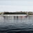 Thumbnail of A waterfront view with buildings, trees, and a large dark-roofed structure reflected in the calm water.